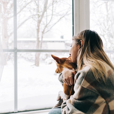 A woman wearing a brown cardigan hugs a small dog while staring out the window. The outdoors is covered in snow.