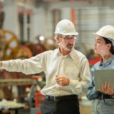 Two engineers, a man and a woman, wearing helmets, inspect heavy machinery during construction installation.