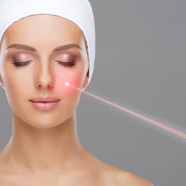A woman with a white towel on her head is getting medical laser treatment on her face with her eyes closed.