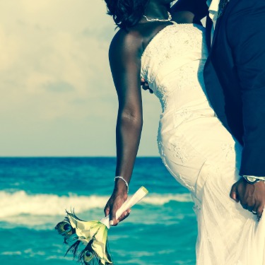 A couple in wedding attire kissing on a beach, with the woman holding flowers and waves in the background.