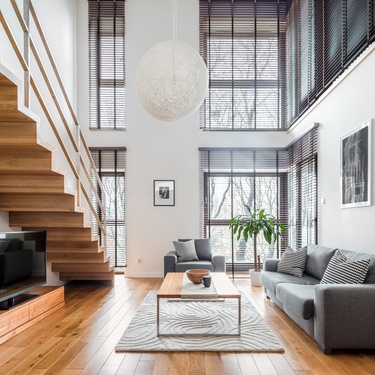 A large living room with a wooden staircase on the left side, wooden floors, multiple couches and large windows.