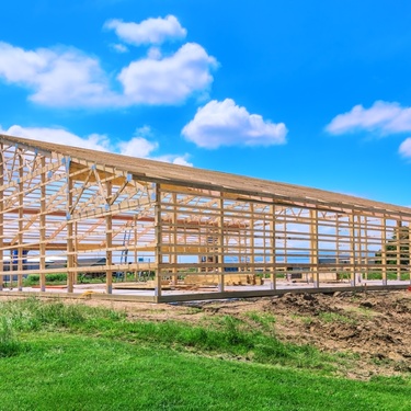 The wooden frame of a large barn on a concrete foundation, surrounding by dirt, green grass, and a bright blue sky.