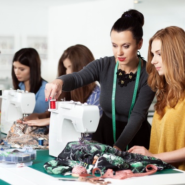 An instructor guiding a student at a sewing machine in a classroom, with other students practicing in the background.
