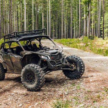 A dark colored UTV sits on a rocky dirt path in a wooded area. The UTV has a black roof rack on top.