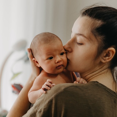 A young mother holding her infant to her chest, supporting their head, and giving them a kiss on the cheek.