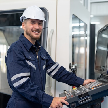 A smiling male technician in uniform and a helmet is operating a CNC machine in an industrial heavy-metal workshop.