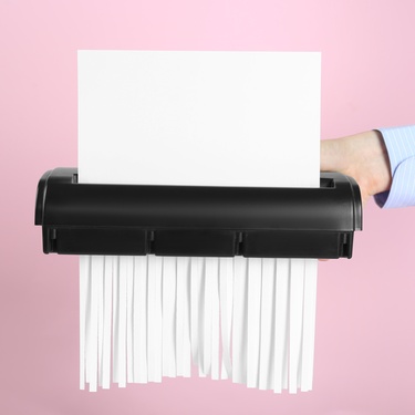 A close-up of someone's hand holding a shredder with paper going through it and shredding into long strips.