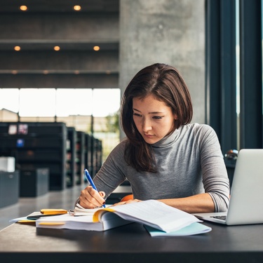 A young woman college student is sitting in the library and is taking notes in her notebook from her textbooks.