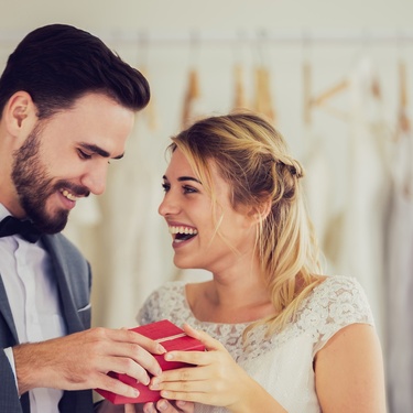 A smiling bride and groom opening a small red gift box together with hanging dresses in the background.