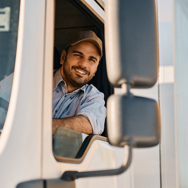 A semi-truck driver smiles from the cab window, wearing a cap and uniform, with hands on the wheel inside the vehicle.