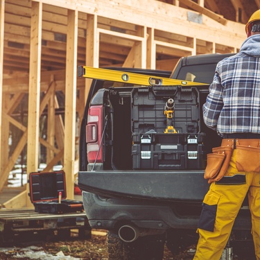 A man wearing a flannel shirt, a helmet, and a tool belt, standing behind a pickup truck at a construction site.