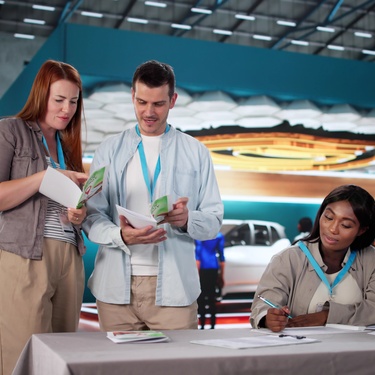 People stand at a trade show, looking at a company's brochures. They are wearing blue lanyards around their necks.