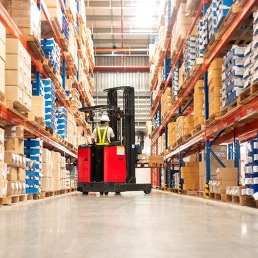 A worker driving a forklift down an aisle of pallet storage systems in a warehouse. The driver is wearing a hard hat.