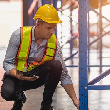 A warehouse engineer checking the safety of the metal rack shelves for inventory. He is holding a tablet.