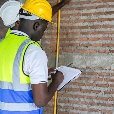 Two site workers wearing hard hats and reflective vests inspect a wall, one taking notes on a clipboard.