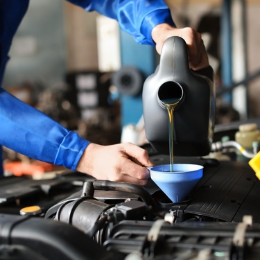 An individual in a blue jumpsuit uses a blue funnel to pour amber-colored liquid from a gray container into a car engine.