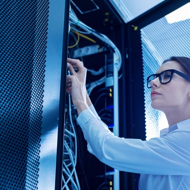 A person with a ponytail wearing eyeglasses and a white long sleeve shirt works with wires in an open service cabinet.