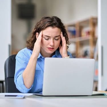 A young woman sits at a white desk with her eyes closed and hands on her temples. An open letter is on the desk.
