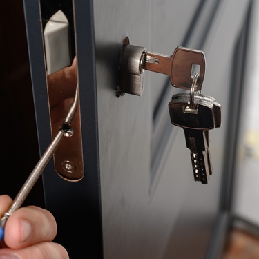 A locksmith uses a tool to install a sleek, silver lock on a black door. The keys are in the keyhole, dangling.