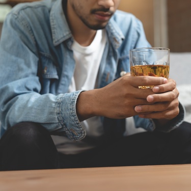 A man sits on a couch holding a glass of whiskey in both hands, looking down with a serious, contemplative expression.