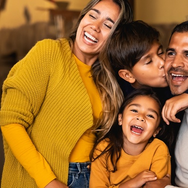 Two adults and two children embracing and smiling enthusiastically. Two of them are wearing matching yellow tops.