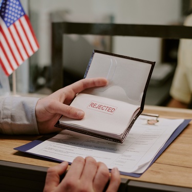 A person holds a passport with the word 'rejected' stamped on it. On the table is a document on a clipboard.