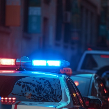 A police car's lights shine brightly in the night, while it is stuck in traffic with multiple cars on the road.