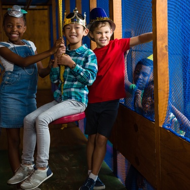 Three children, each wearing a fun costume hat, stand in the corridor of an indoor playground. The child in the middle sits on a swing.
