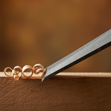 A woodworking chisel carves the edge of a piece of wood. It creates a small curled strip of wood as it moves.
