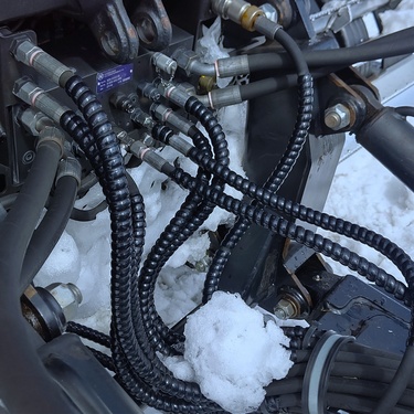 A close-up of a part of a hydraulic system on some machinery. It features tubes and fitings, some of which are covered in snow.