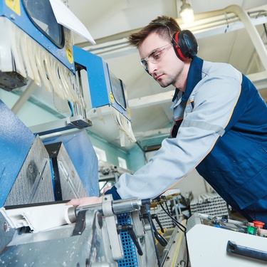 A worker wearing a blue uniform and noise-cancelling headphones, touching a large machine inside a warehouse.