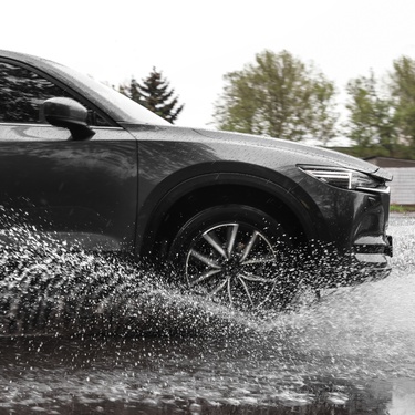 The front of a car driving through a large puddle on a rainy day. The puddle of water is splashing upwards.