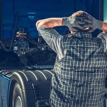 A stressed truck driver standing with his hands behind his head, facing the blue tractor of his semitruck.