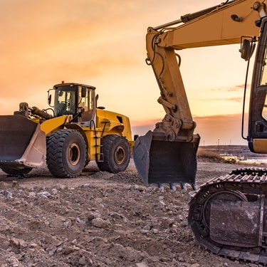The sun sets in the horizon as two yellow excavators work on a construction site filled with rocks.