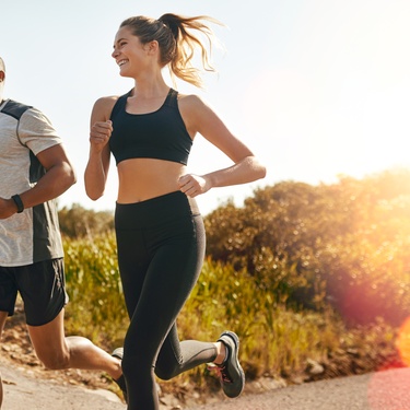 A man and a woman in exercise clothing are smiling while running outside with the sun low in the sky.