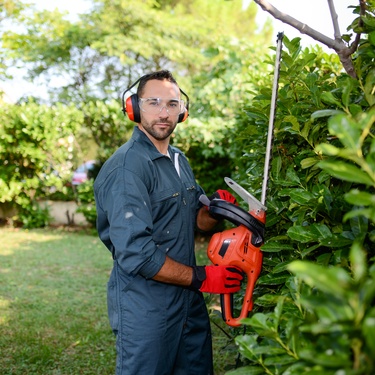 A landscaper wearing a jumpsuit, safety glasses, hearing protection, and gloves using a trimmer to cut hedges.