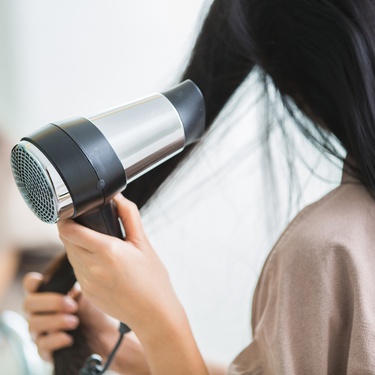 A person in a brown shirt using a silver and black blow dryer to dry their long, straight dark hair.