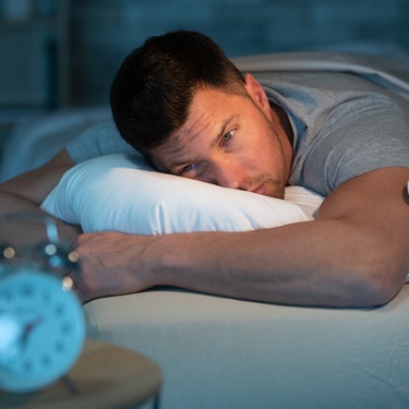 A man lies awake in bed, his head on a pillow, unable to sleep. An alarm clock sits nearby on the night table.