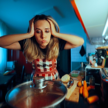 A stressed woman with her hands to her head looks at a pot of soup on the stove next to a cutting board full of ingredients.