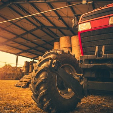 A red tractor parked in a large storage area on a farm. The storage room is full of hay and wooden pallets.