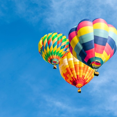 Three rainbow colored hot air balloons floating in the sky. The sky is clear and blue with no clouds.