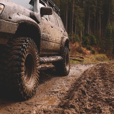 An SUV is parked on a muddy trail in the woods. The vehicle has mud splattered on the body and tires.