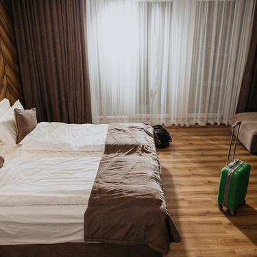 Cozy hotel room with neatly made bed, wooden accents, large window with sheer curtains, and green suitcase on floor.