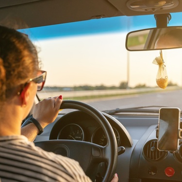A woman is wearing sunglasses and driving a vehicle as the sun shines through the windshield and reflects off the interior.