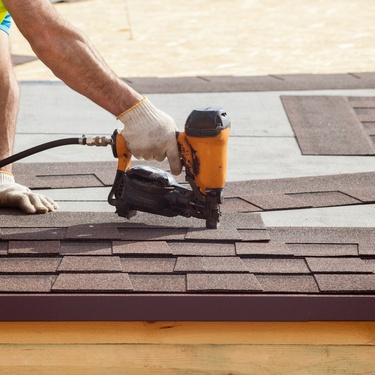 An individual in a yellow safety vest and white gloves using an orange nail gun to install shingles on a roof.