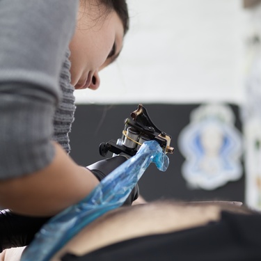 A close-up shows a woman wearing a gray shirt using a black and gray tattoo machine to apply a tattoo on a customer.