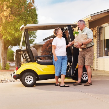 A senior man and woman standing in the driveway next to a yellow golf cart after using it to get groceries.