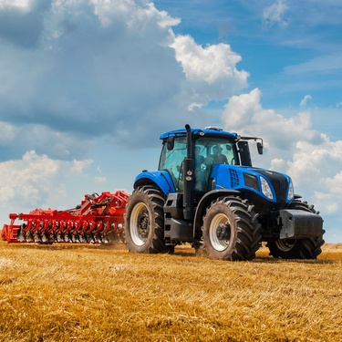A brand-new blue tractor with a red harrow operates in a large grassy field beneath a partly cloudy sky.