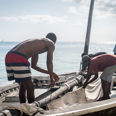 A group of Black men is working on a boat, helping to set up the cloth-sailing flag, while wearing swimming trunks.