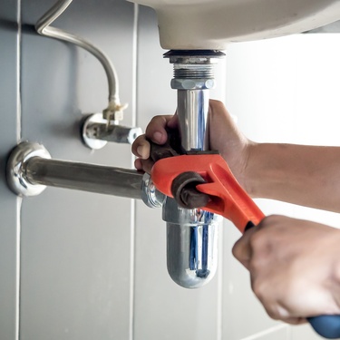 A close-up shows a person using a black and orange wrench to tighten exposed silver water pipes under a white sink.